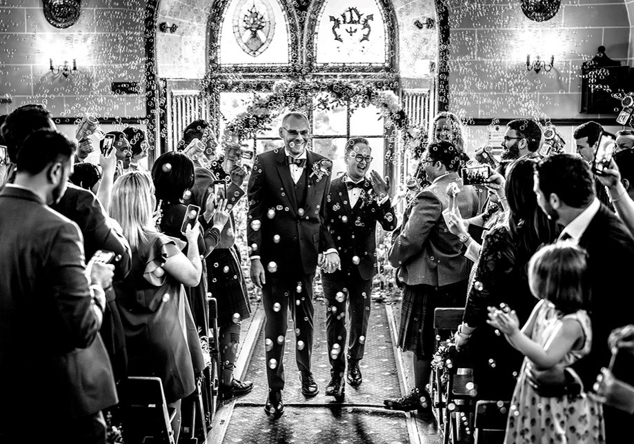 Two smiling grooms walk back down aisle while guests throw confetti